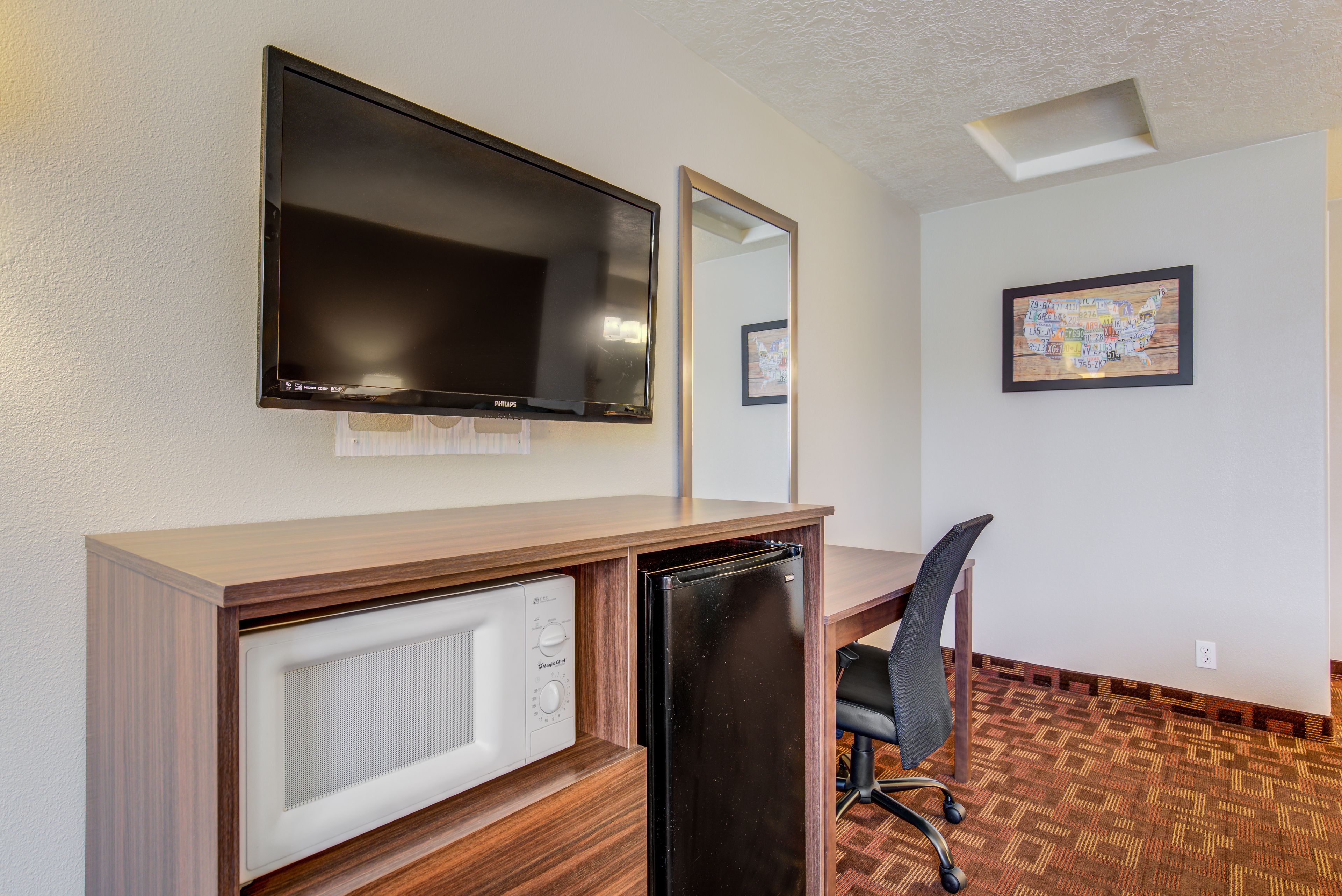 A hotel room with a wooden desk, a black refrigerator, a microwave, a television, a chair, and a framed map on the wall.