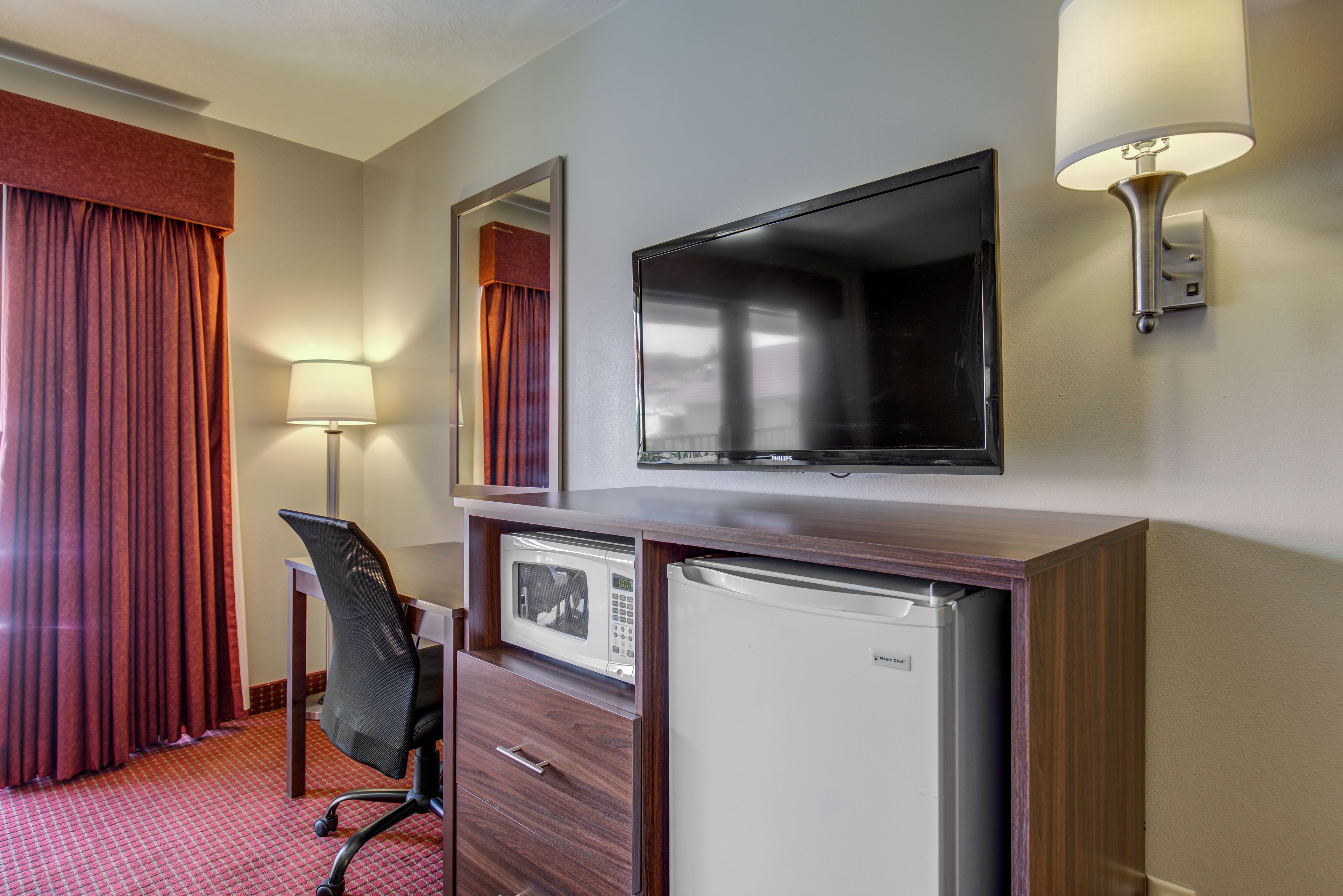 A hotel room with a flat-screen TV, a microwave, a refrigerator, a desk with a chair, and a lamp.
