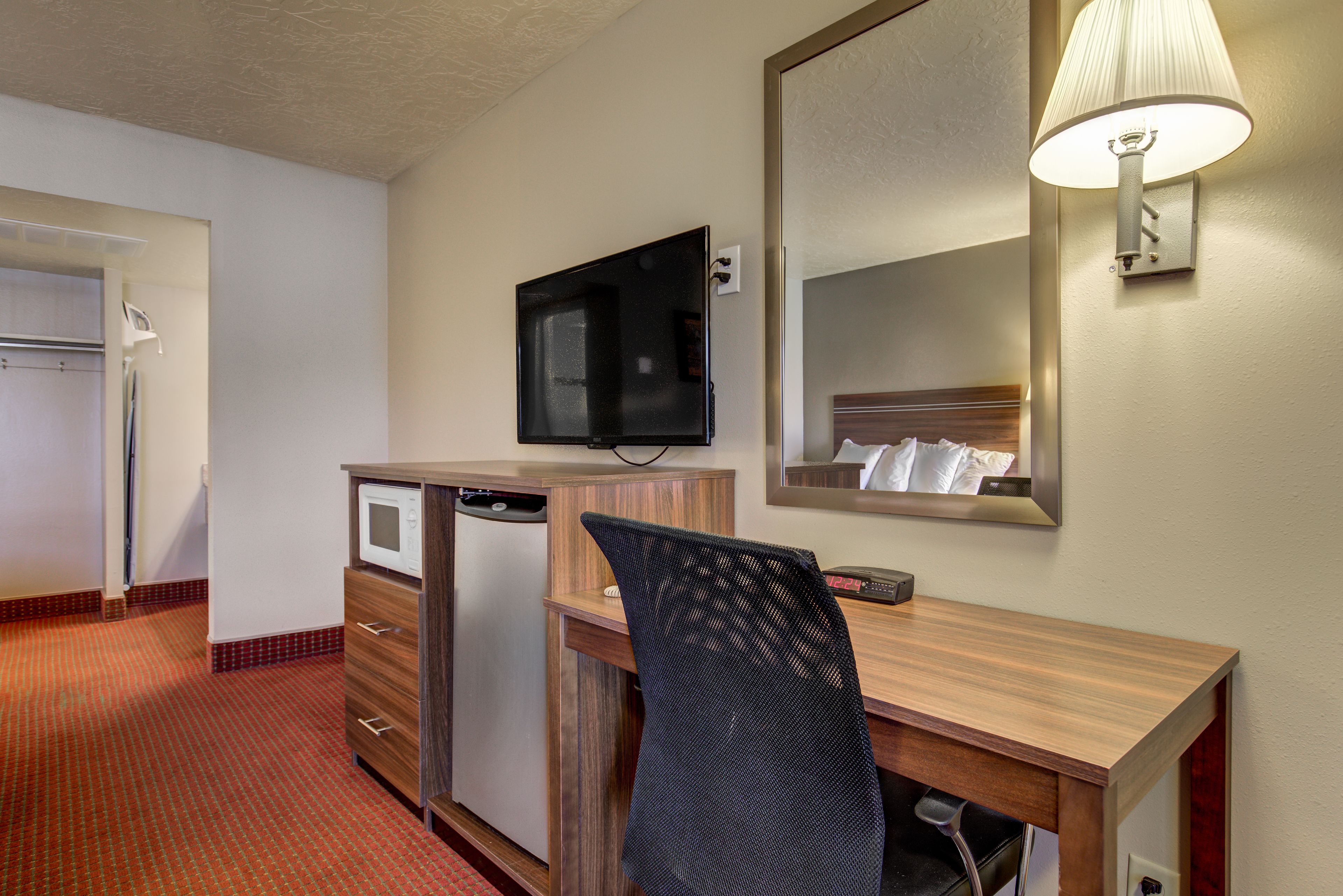 A hotel room with a wooden desk, a microwave, a refrigerator, a television, and a mirror with a lamp.