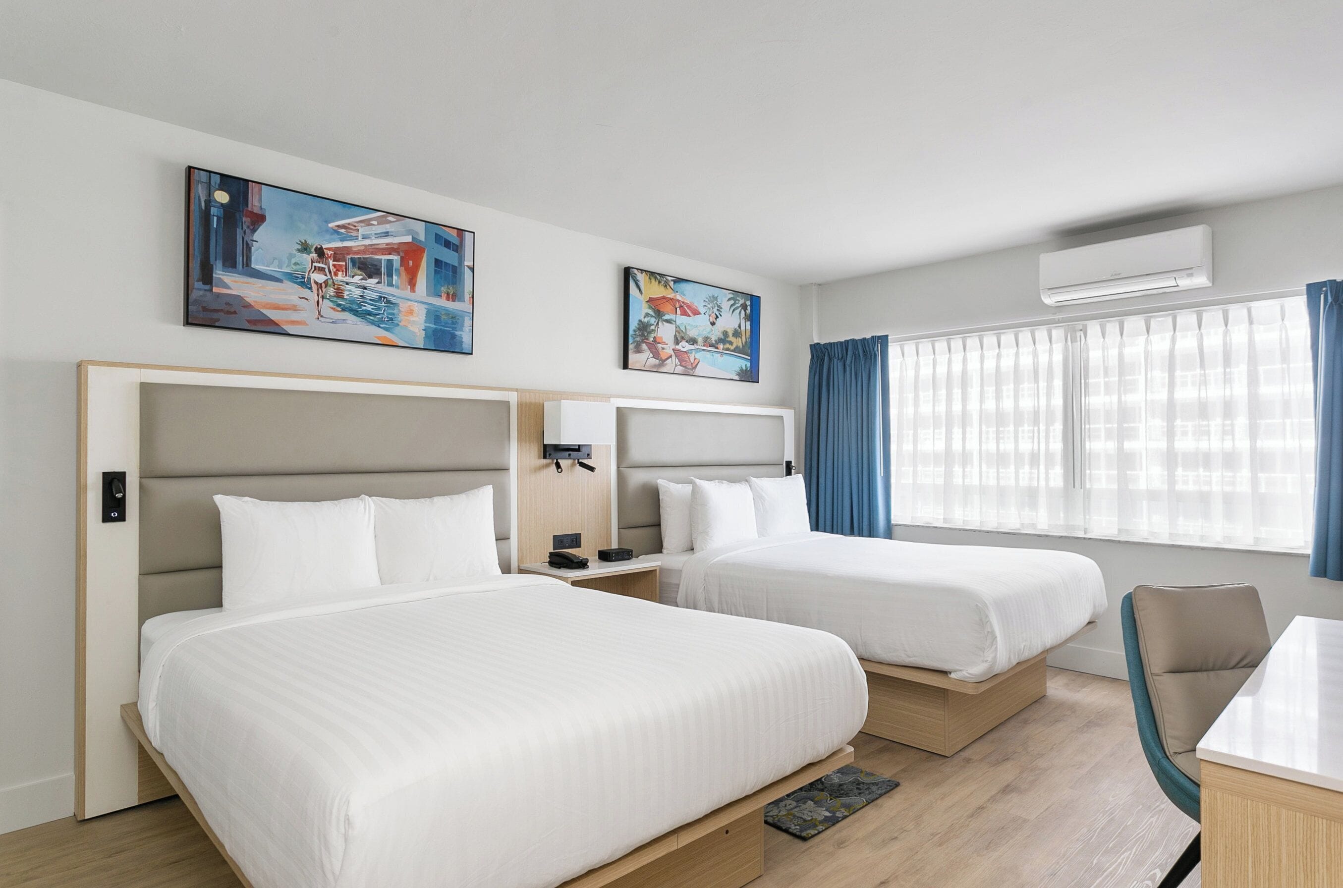 Habitación de hotel con dos camas, un escritorio y una silla. Hay cuadros en la pared y un equipo de aire acondicionado sobre la ventana.