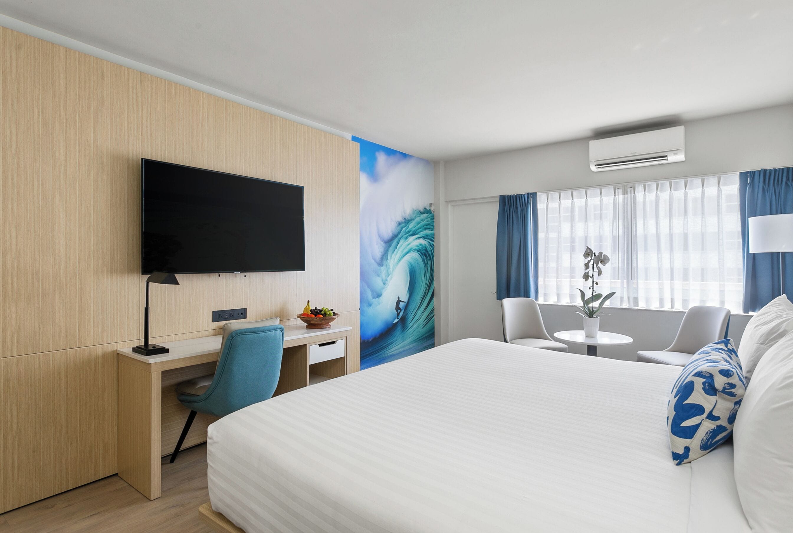 A hotel room with a bed, a desk with a chair, a TV, a wall mural, a small table with a plant, and a window with curtains.