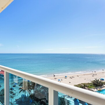 A balcony with a view of the ocean and beach, glass railings, and outdoor seating.