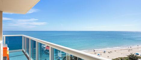 A balcony with a view of the ocean and beach, glass railings, and outdoor seating.
