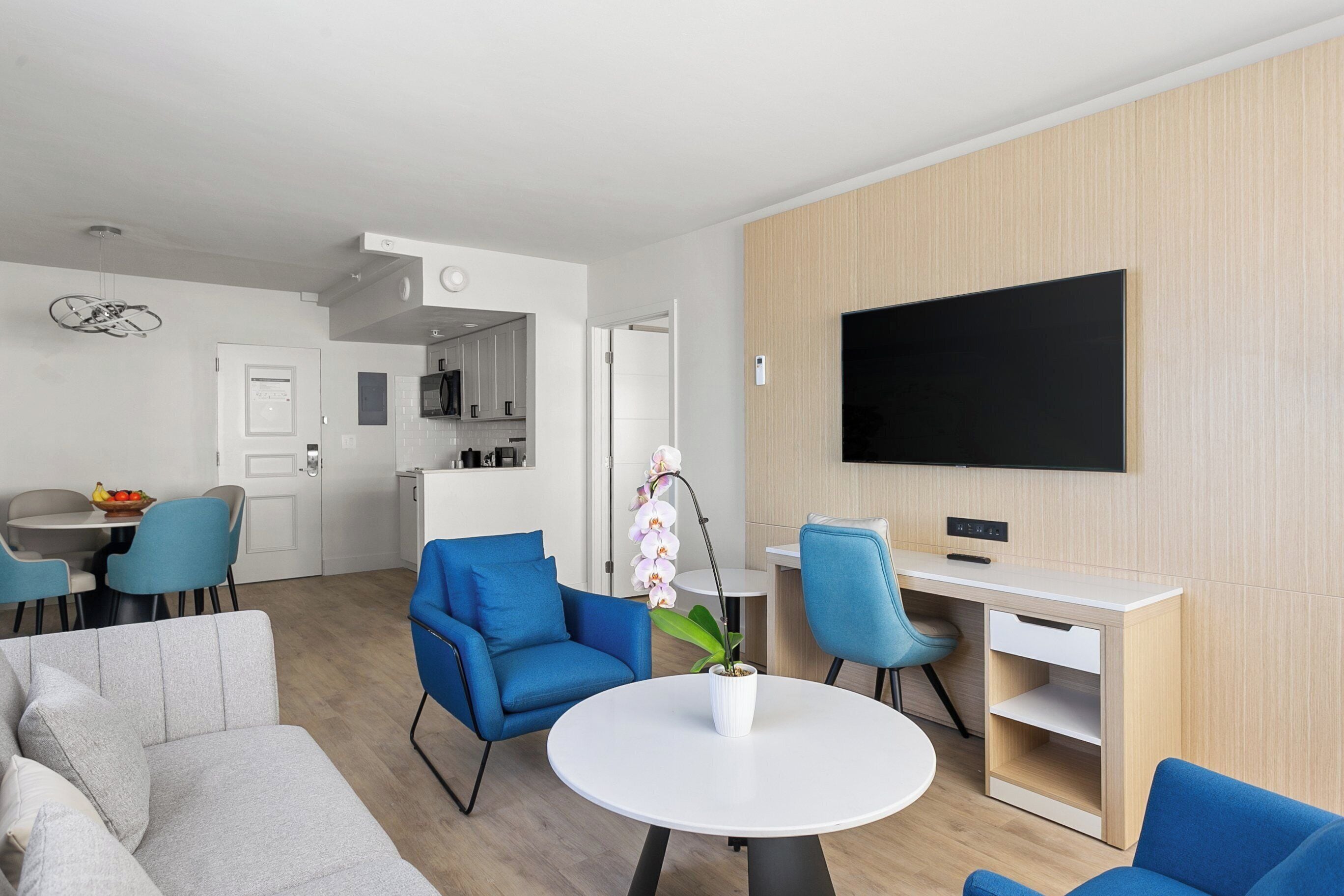 Un salon moderne avec un canapé gris, des fauteuils bleus, une table basse blanche et un téléviseur à écran plat fixé au mur.