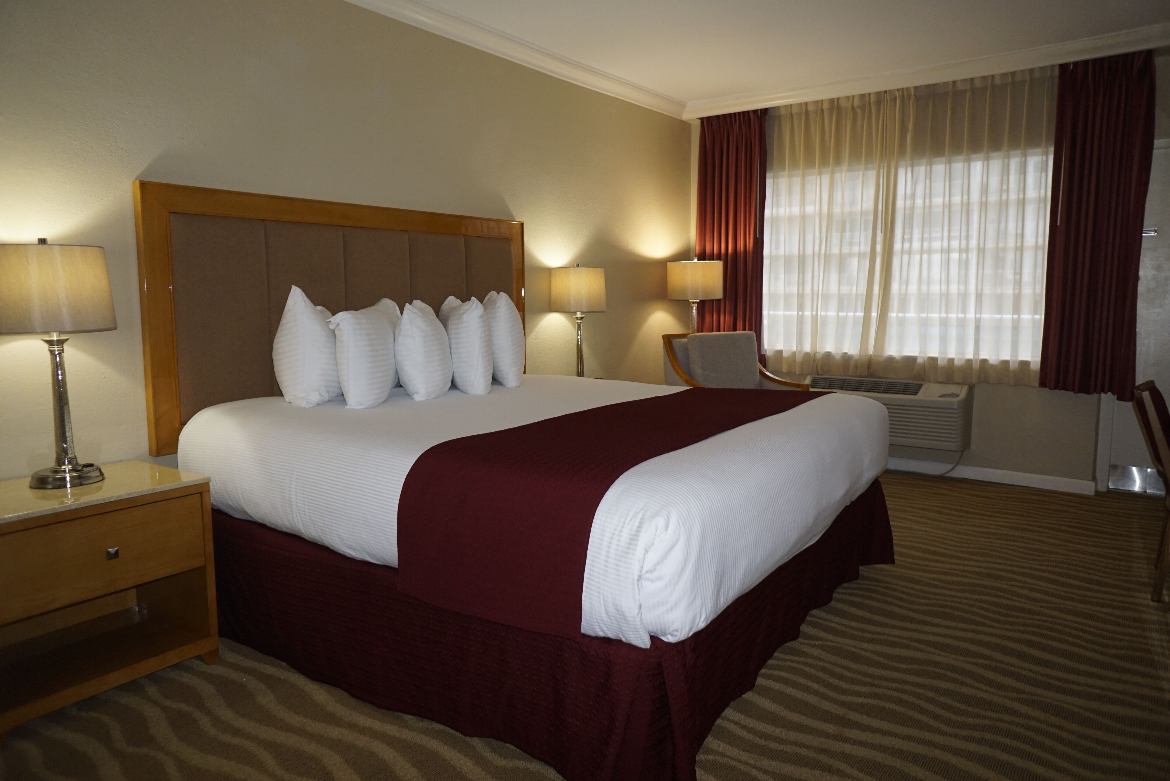A hotel room with a large bed, two bedside lamps, an air conditioning unit, a desk, and a chair.