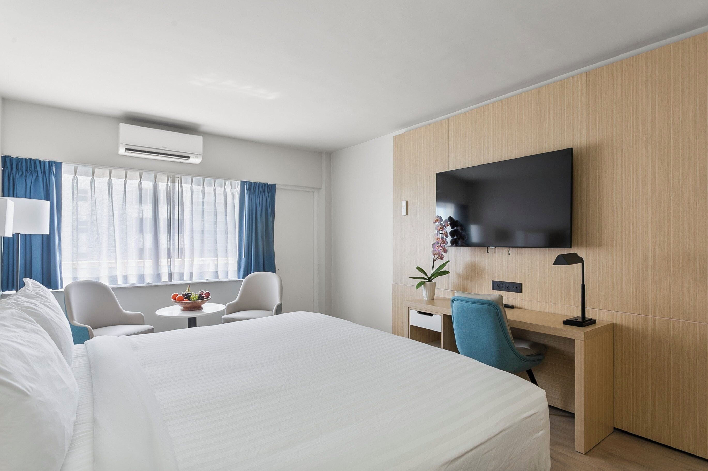 A hotel room with a large bed, a desk with a chair, a TV mounted on the wall, and a small table with a plant.