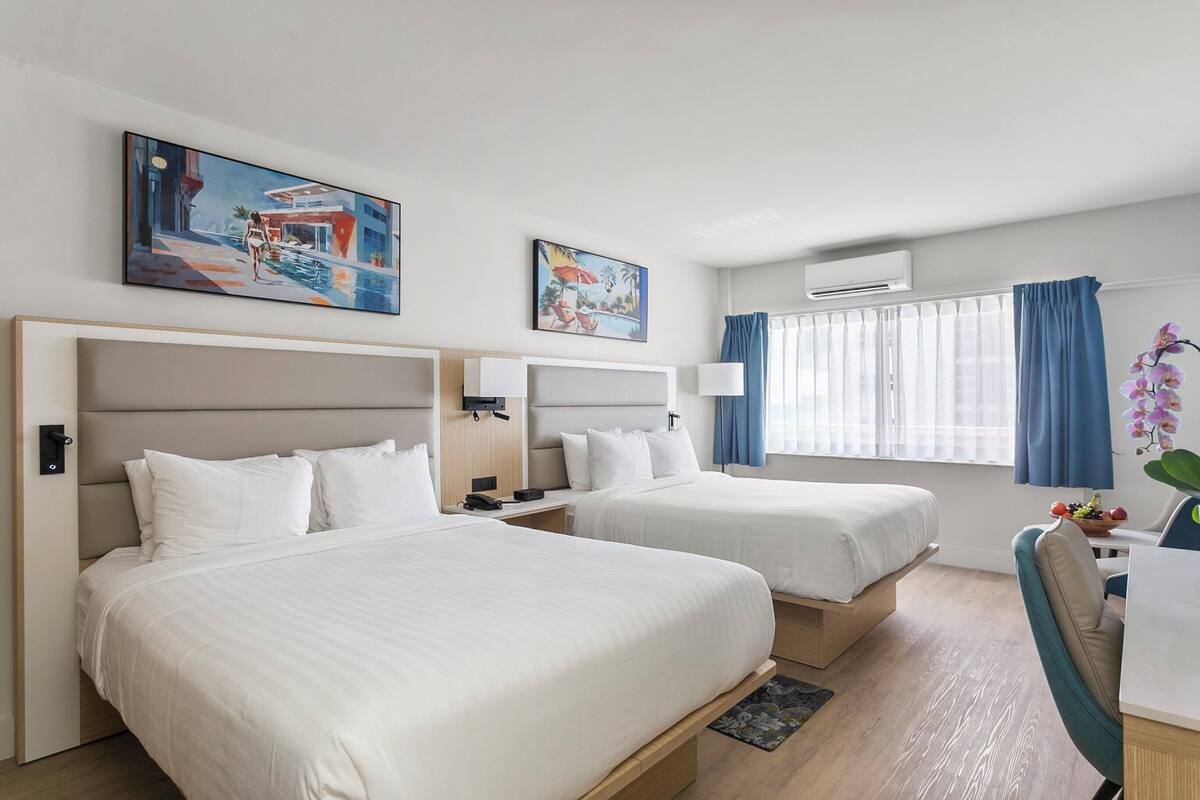 A hotel room with two beds, a desk with a chair, a window with curtains, and artwork on the walls.
