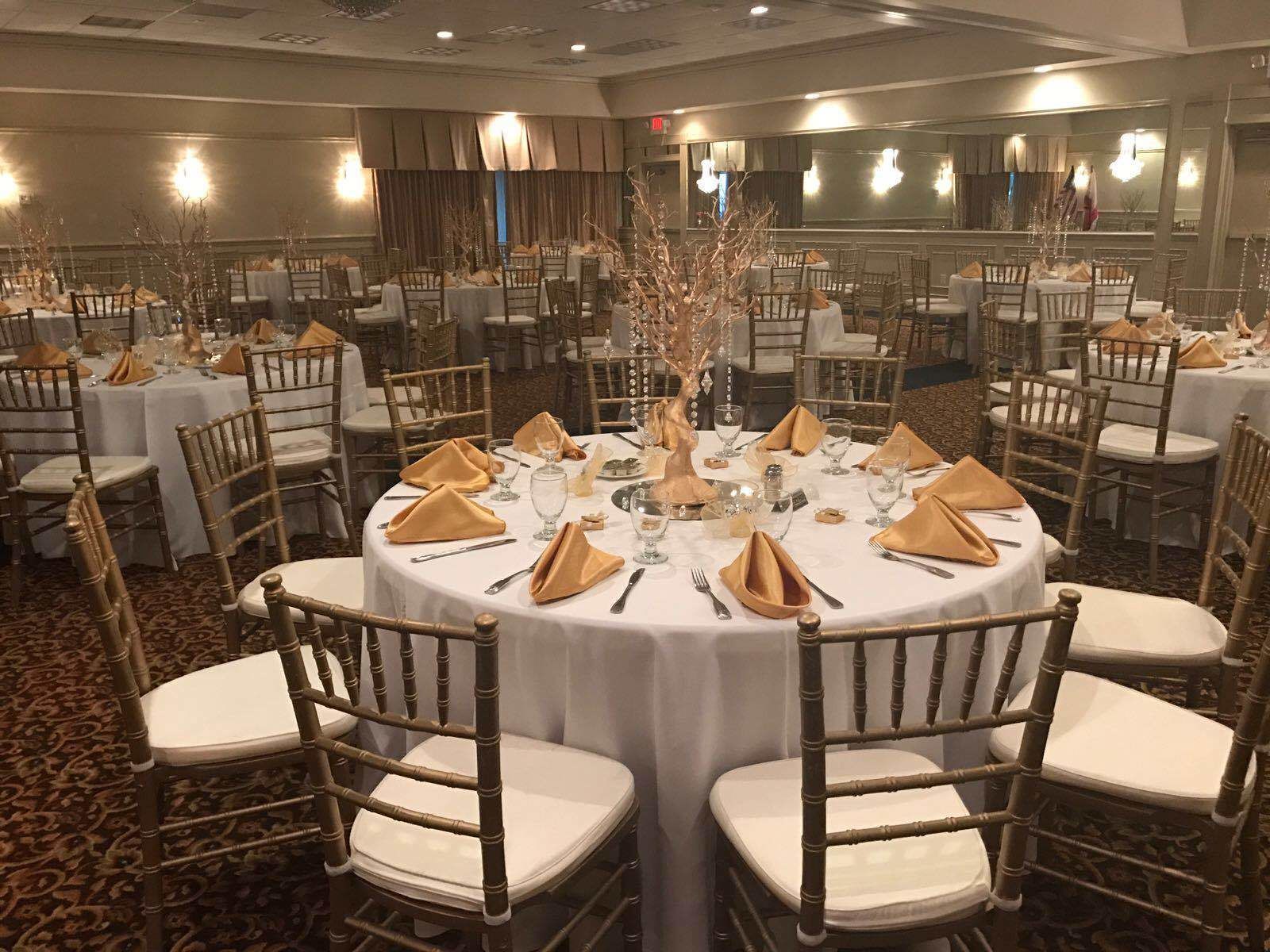 Un salón de banquetes con mesas redondas dispuestas para comer, cada una con manteles blancos, servilletas doradas y sillas de madera.