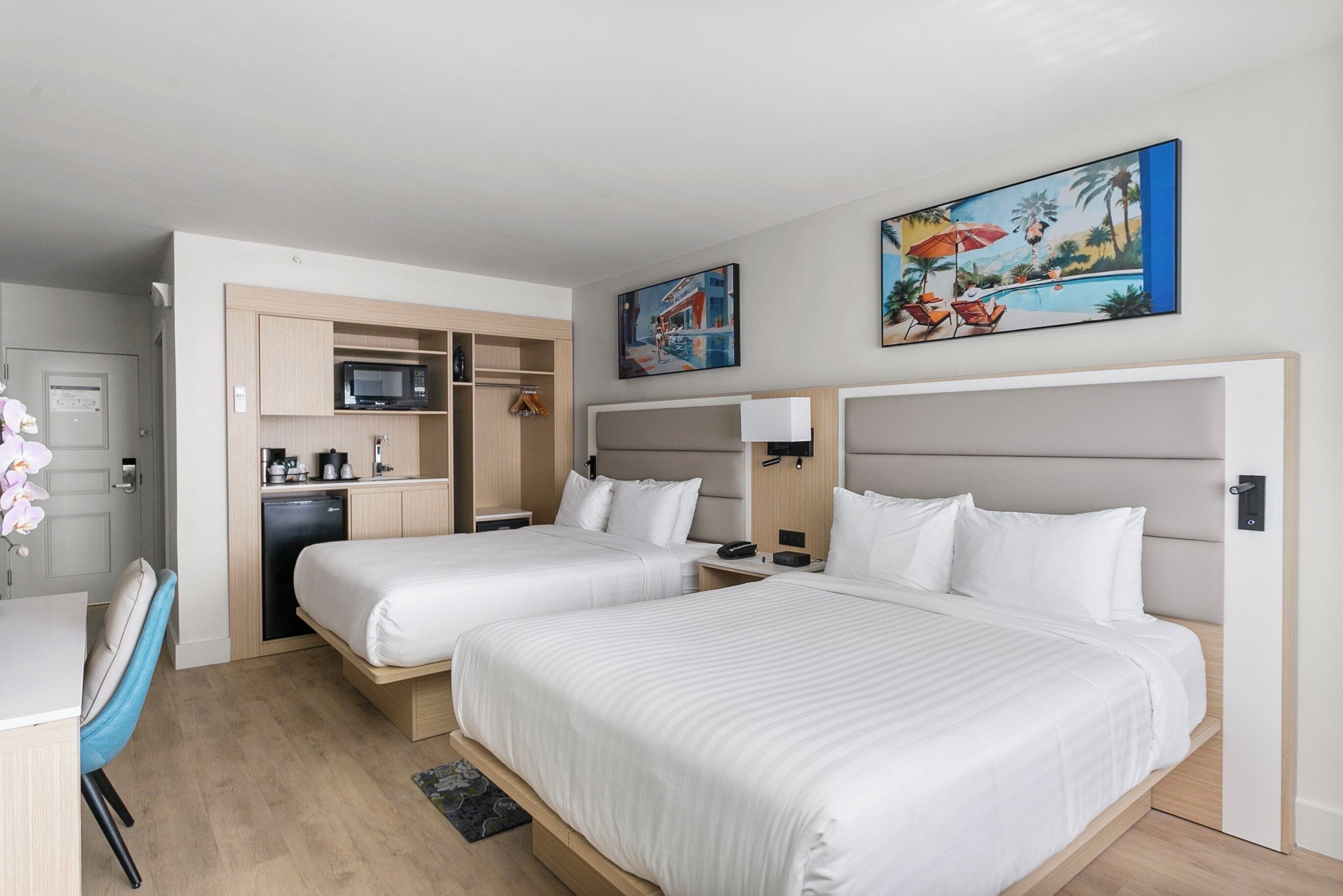A hotel room with two beds, a desk, a kitchenette, and artwork on the walls.