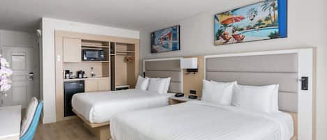 A hotel room with two beds, a desk, a kitchenette, and artwork on the walls.