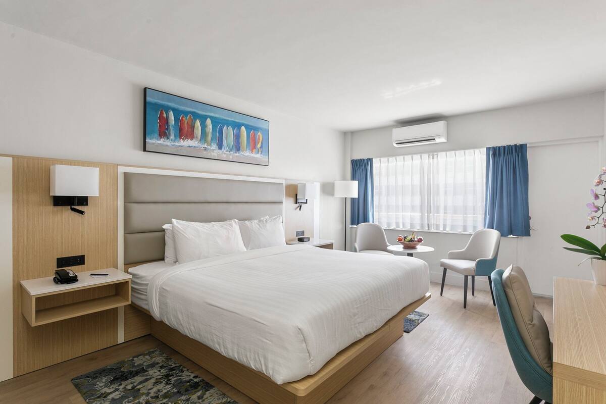 A hotel room with a large bed, bedside tables, a desk with a chair, a window with curtains, an air conditioning unit, and a painting on the wall.