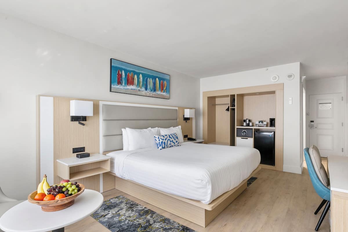 A hotel room with a bed, bedside tables, a small desk, a chair, a fruit bowl, a painting, and a kitchenette.