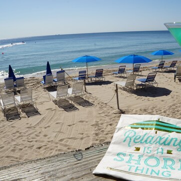 A beach with rows of blue umbrellas and lounge chairs, a clear view of the ocean, and a towel with the text "Relaxing is a shore thing".