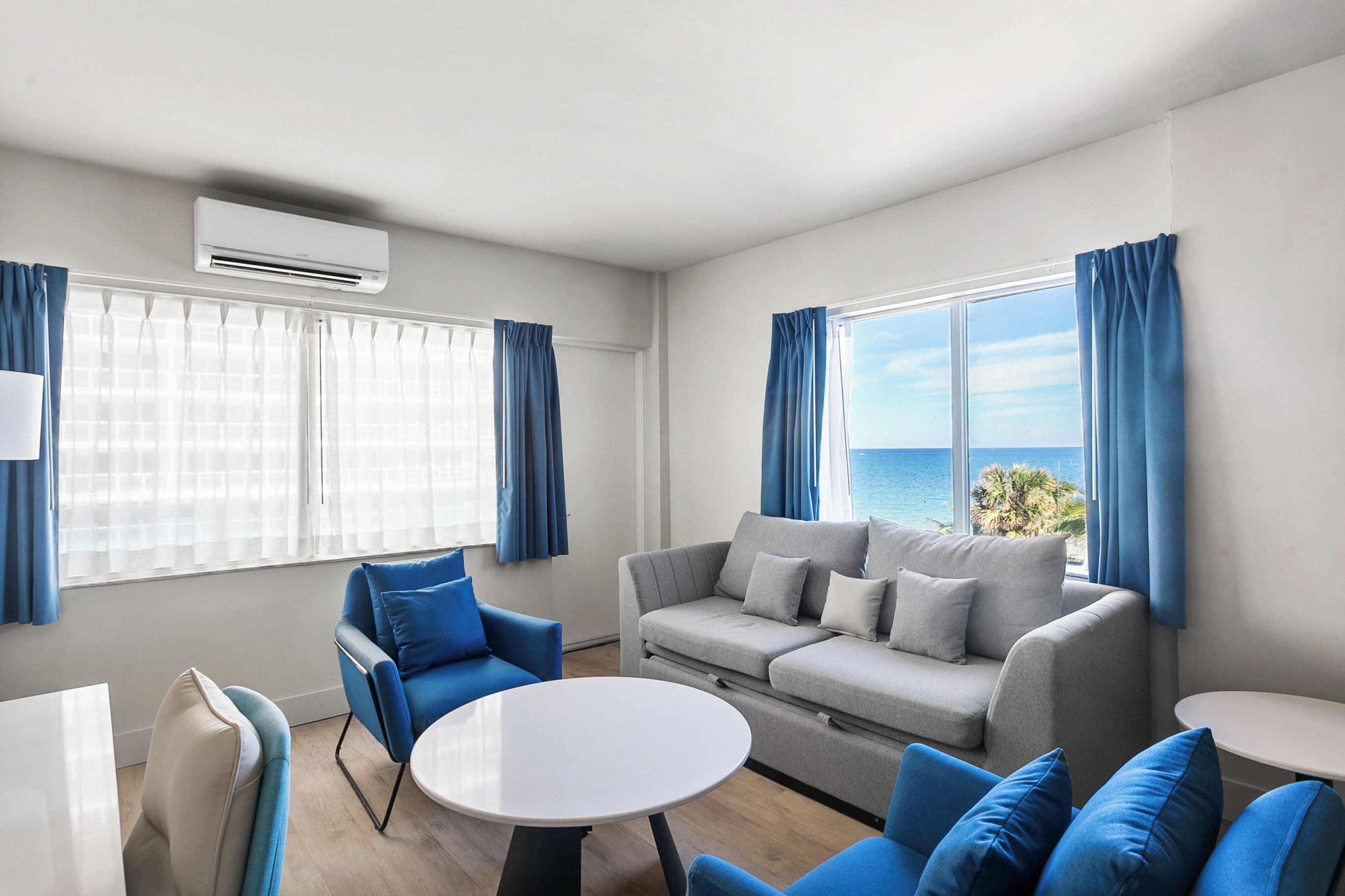 A modern living room with a grey sofa, blue armchairs, a white coffee table, and a view of the ocean.