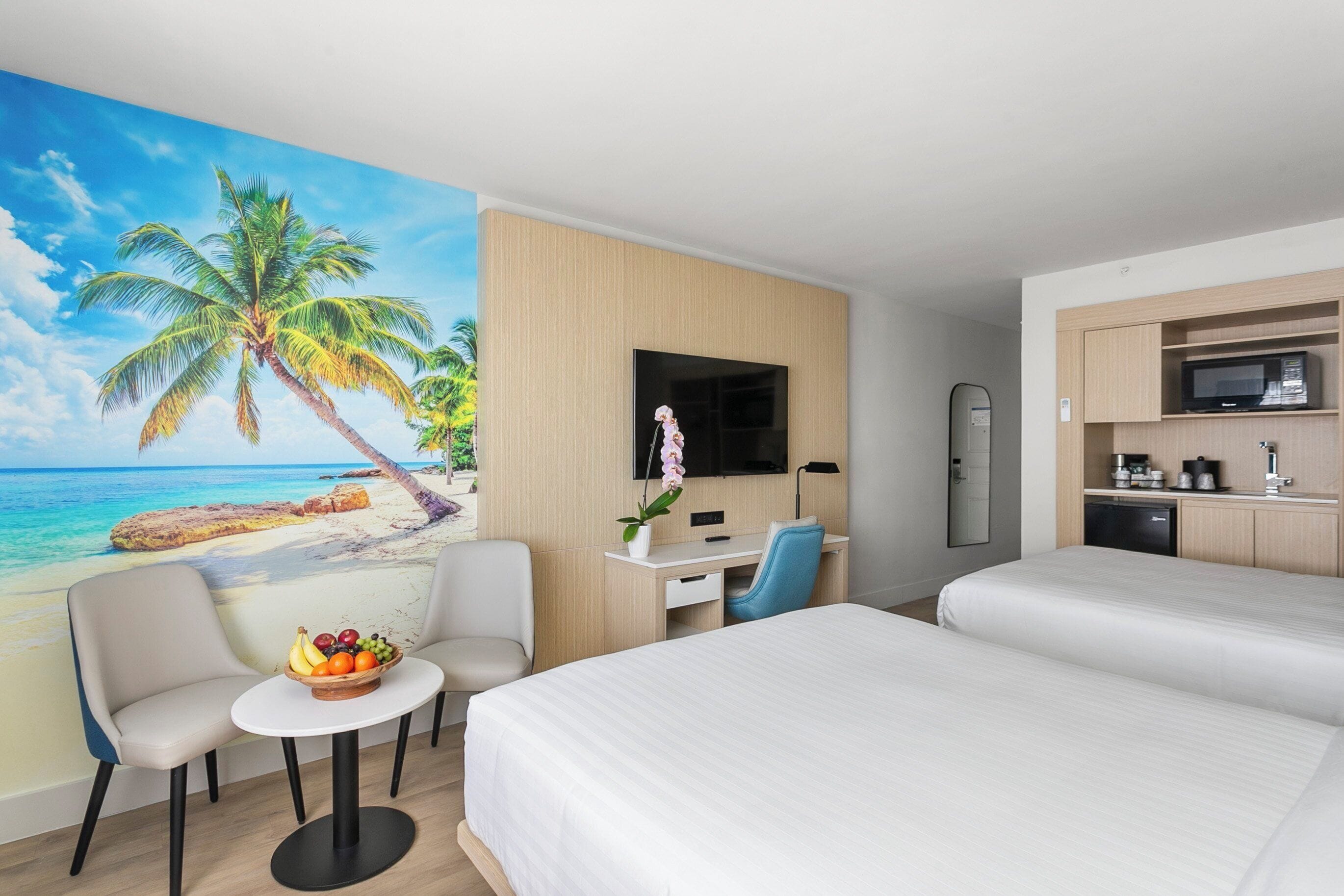 A hotel room with a beach mural, a desk with a chair, a bed, and a small table with fruit.