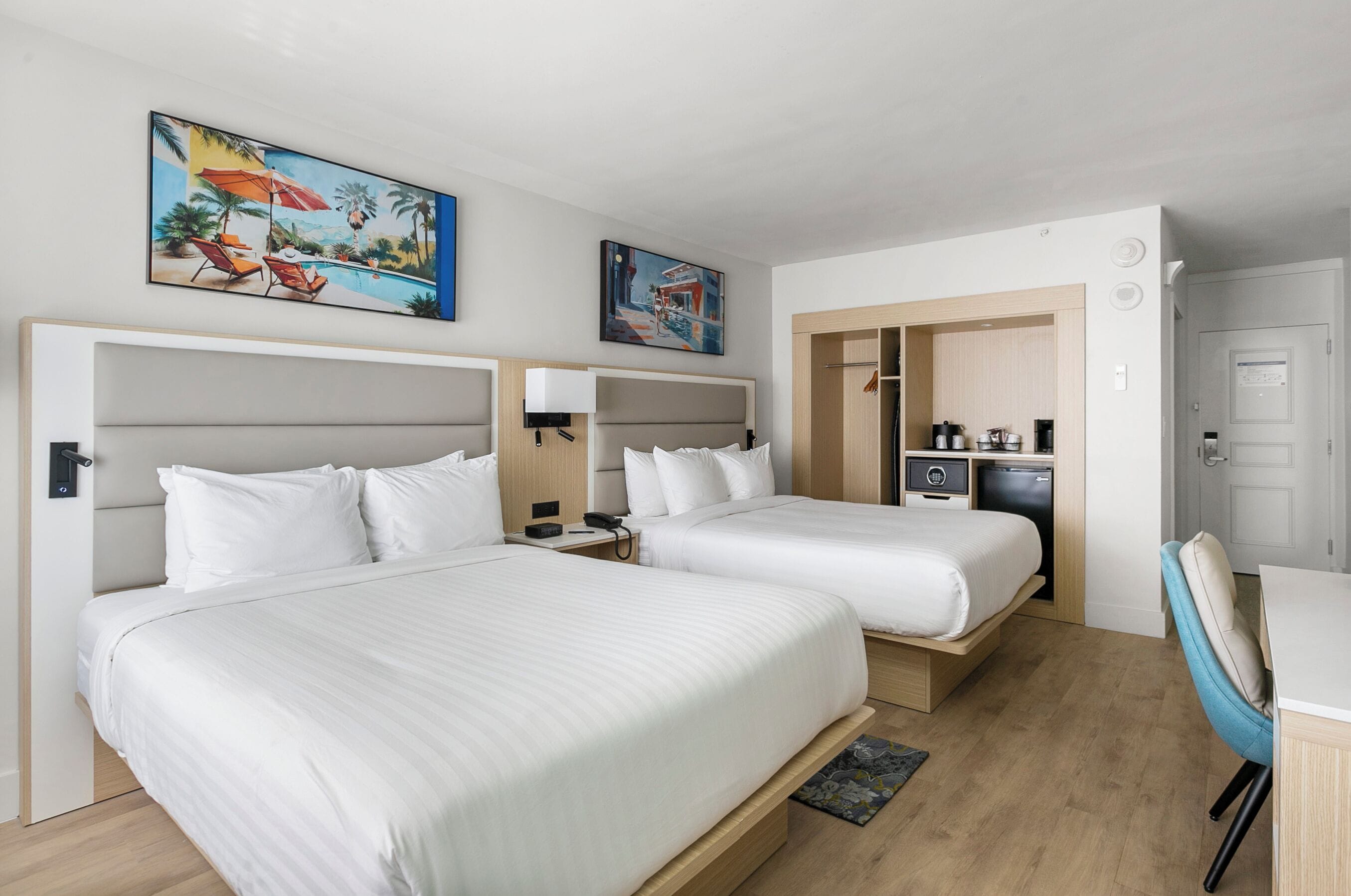 A hotel room with two beds, a desk, a chair, a small kitchenette, and artwork on the walls.