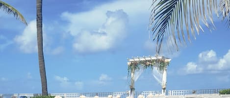 A beach wedding setup with white chairs and a floral arch.