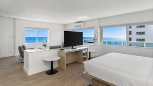 A modern hotel room with a large bed, a desk with a TV, a small table, and a view of the ocean.