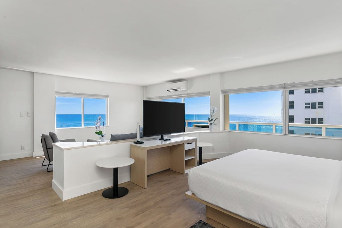 A modern hotel room with a large bed, a desk with a TV, a small table, and a view of the ocean.