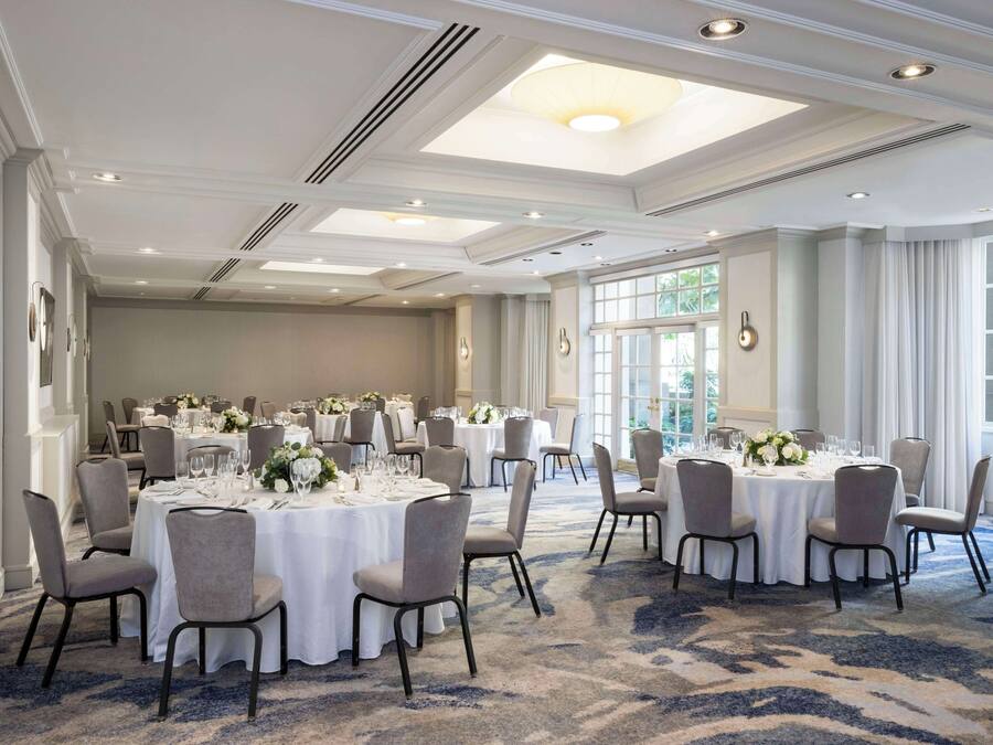 A spacious banquet hall with round tables set for dining, large windows, and a patterned carpet.