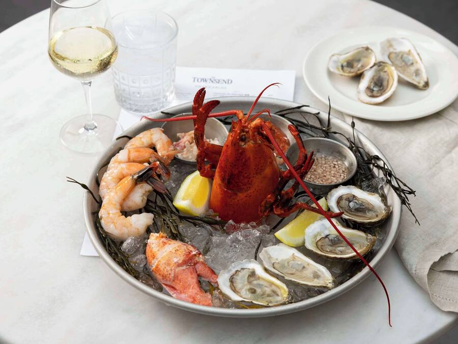 A seafood platter with shrimp, lobster, and oysters on ice, accompanied by a glass of white wine and a menu.