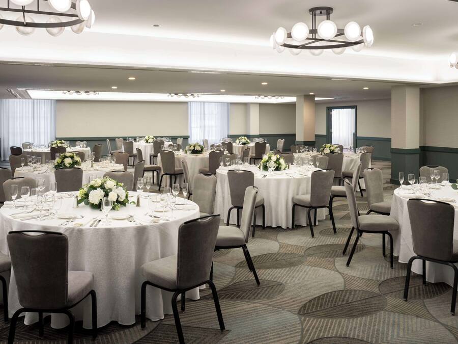 Un salón de banquetes con mesas redondas dispuestas para comer, cada una con mantel blanco y centros de mesa florales.