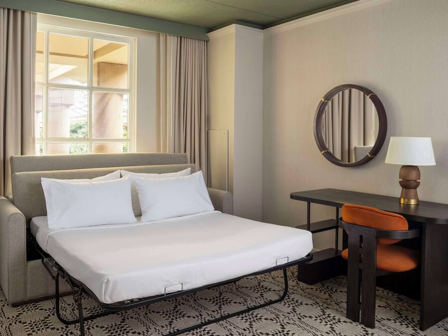A hotel room with a bed, a desk, a chair, a lamp, and a window with curtains.