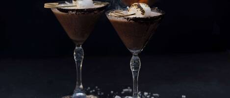 Dos copas de martini con licor de chocolate, un fósforo encendido y un malvavisco sobre una superficie oscura.