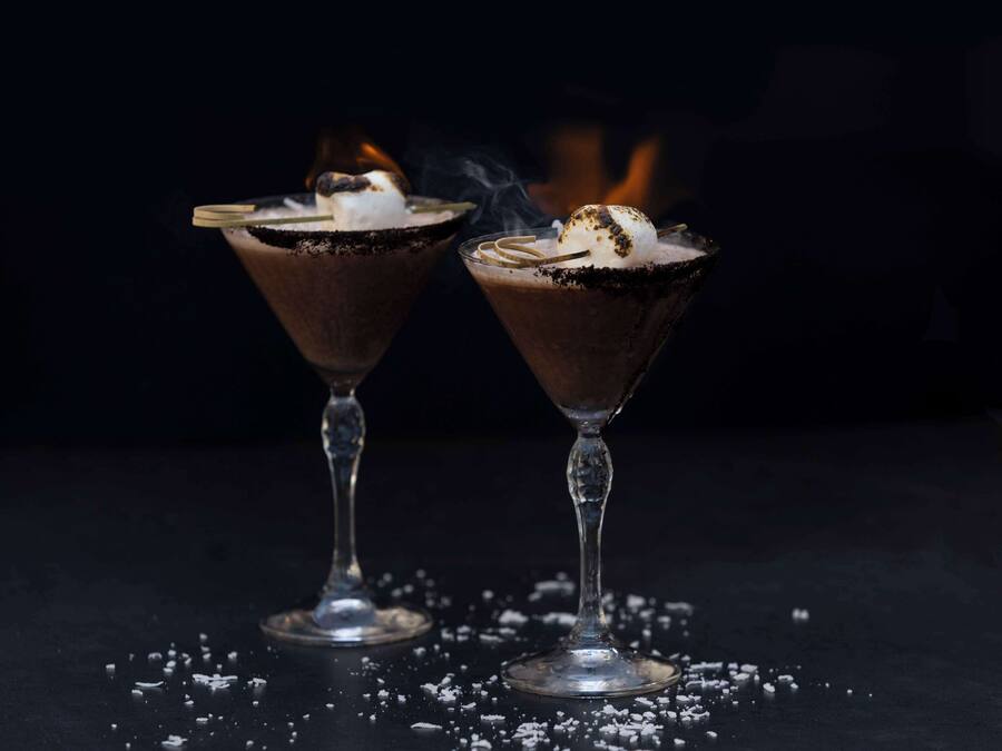 Dos copas de martini con licor de chocolate, un fósforo encendido y un malvavisco sobre una superficie oscura.