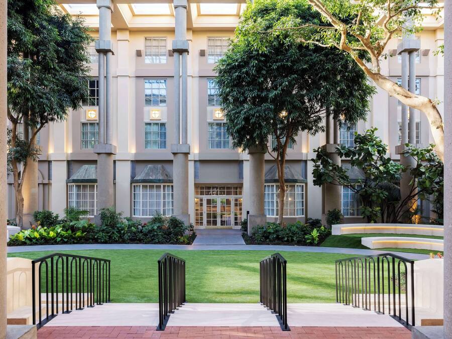 A courtyard with a central pathway, surrounded by greenery and a building with large windows and columns.