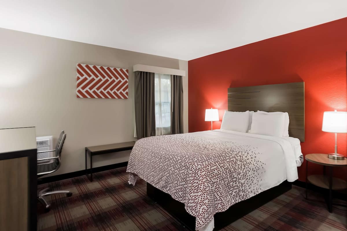 A hotel room with a bed, a desk, a chair, a lamp, and a window with curtains.