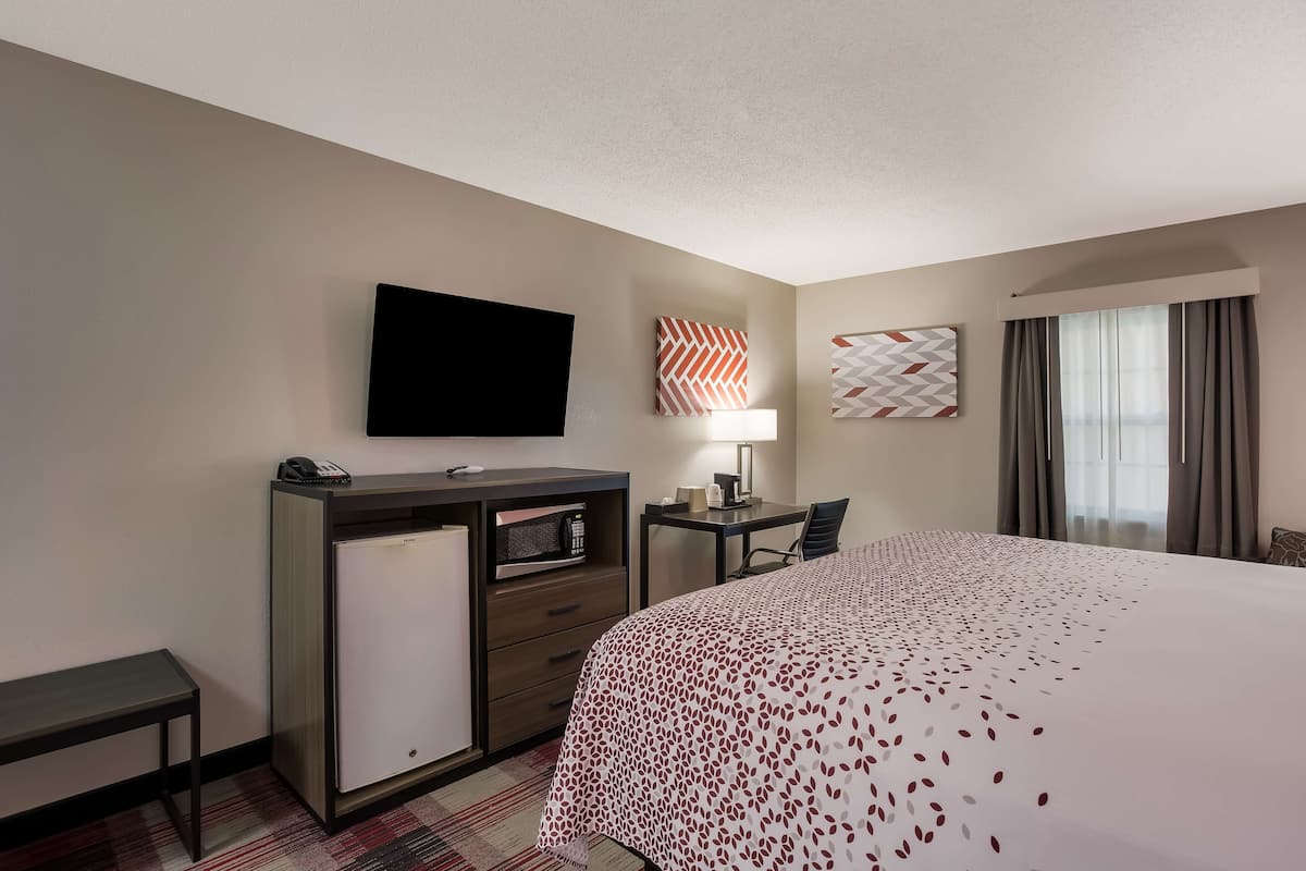 A hotel room with a bed, a desk with a lamp, a flat-screen TV, a minibar, and a window with curtains.