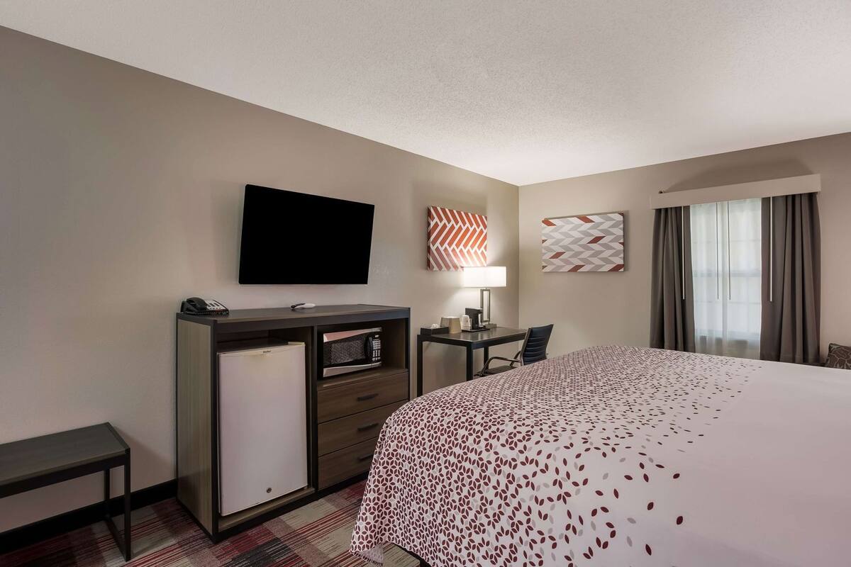 A hotel room with a bed, a desk, a flat-screen TV, a minibar, and a window with curtains.