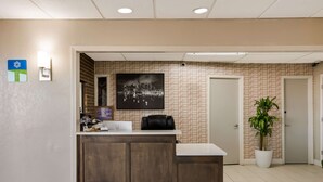 A reception area with a wooden reception desk, a cityscape painting, a potted plant, and a door in the background. - SureStay Hotel by Best Western Higginsville (Higginsville)