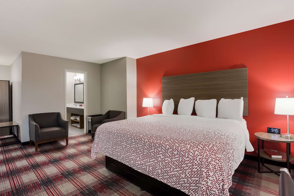 A hotel room with a large bed, a desk with a chair, a small table with a lamp, and a red accent wall.