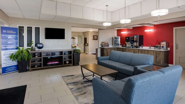 A modern living room with a blue sofa, a coffee table, and a fireplace. There is a kitchen area with a red accent wall and a flat-screen TV mounted above the counter. - SureStay Hotel by Best Western Higginsville (Higginsville)