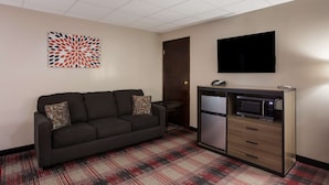 A modern hotel room with a dark sofa, a patterned rug, a flat-screen TV, and a minibar. - SureStay Hotel by Best Western Higginsville (Higginsville)