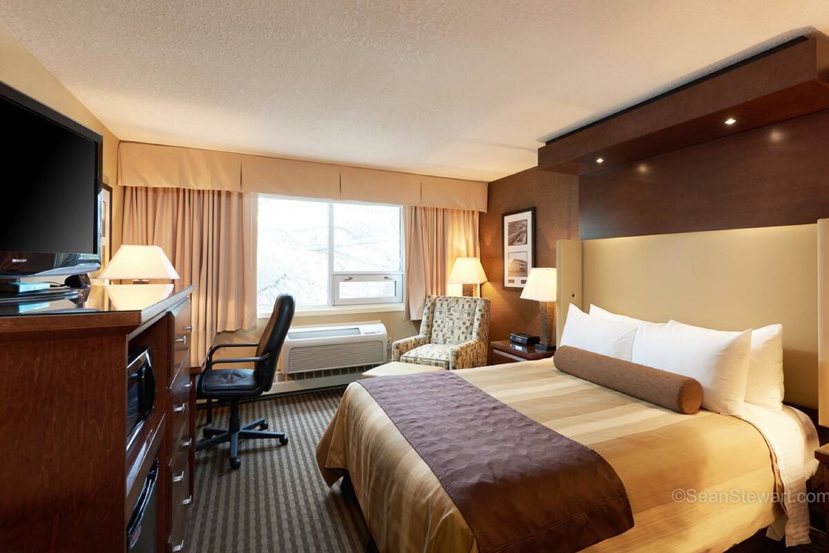 Ein Hotelzimmer mit einem großen Bett, einem Schreibtisch mit Fernseher, einem Sessel und einem Fenster mit Vorhängen.