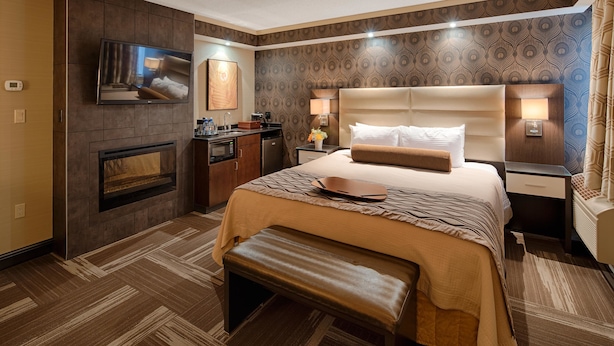 Habitación de hotel con una cama grande, un televisor de pantalla plana en la pared, una kitchenette pequeña y un área de descanso con un banco.