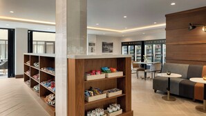 Un interior moderno con estantes de madera que exhiben aperitivos, una zona de estar con sofá y mesas, y un refrigerador identificado con el rótulo 'The Market' al fondo.