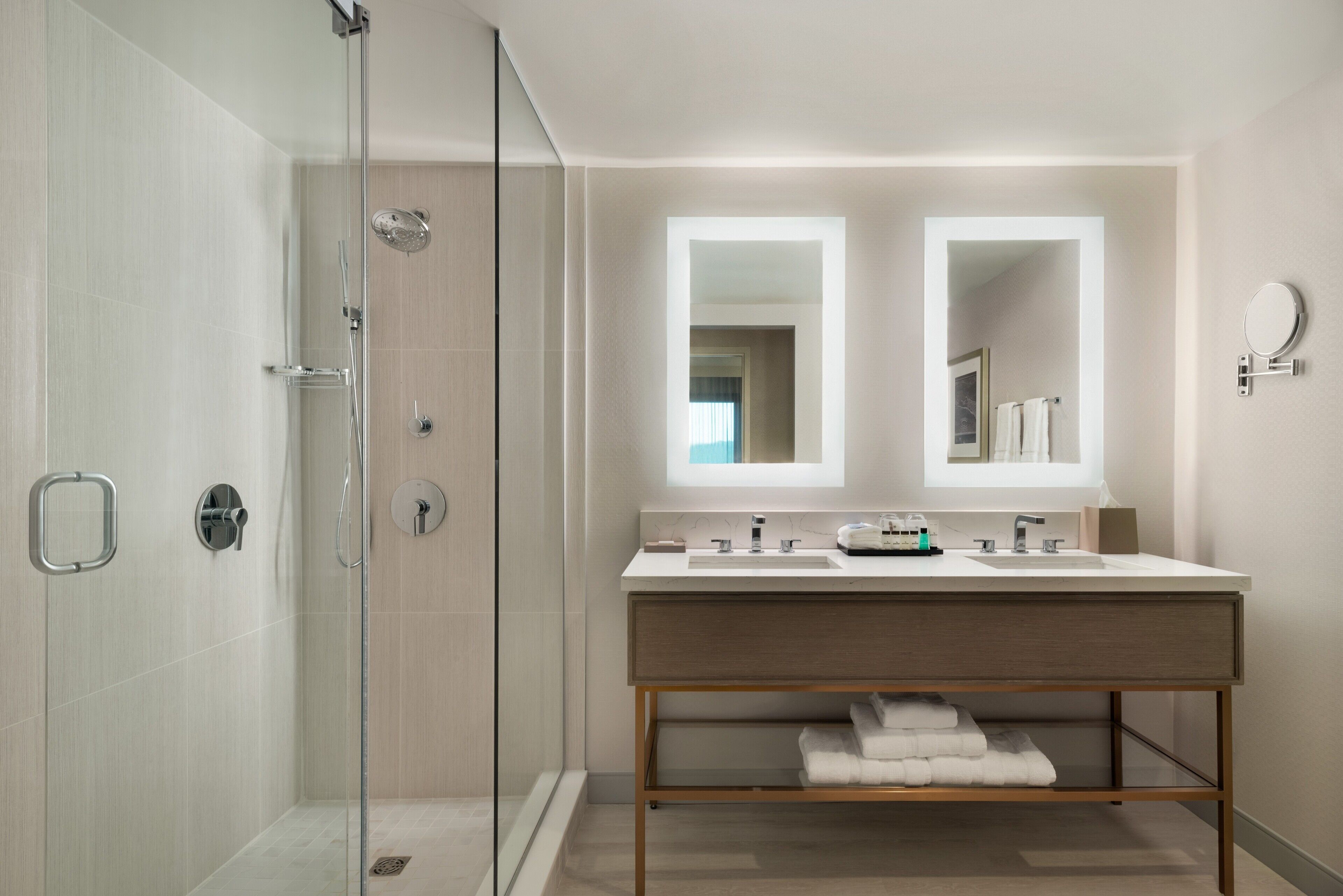 A modern bathroom with a glass-enclosed shower, a double vanity with two mirrors, and a wooden vanity with a towel rack.