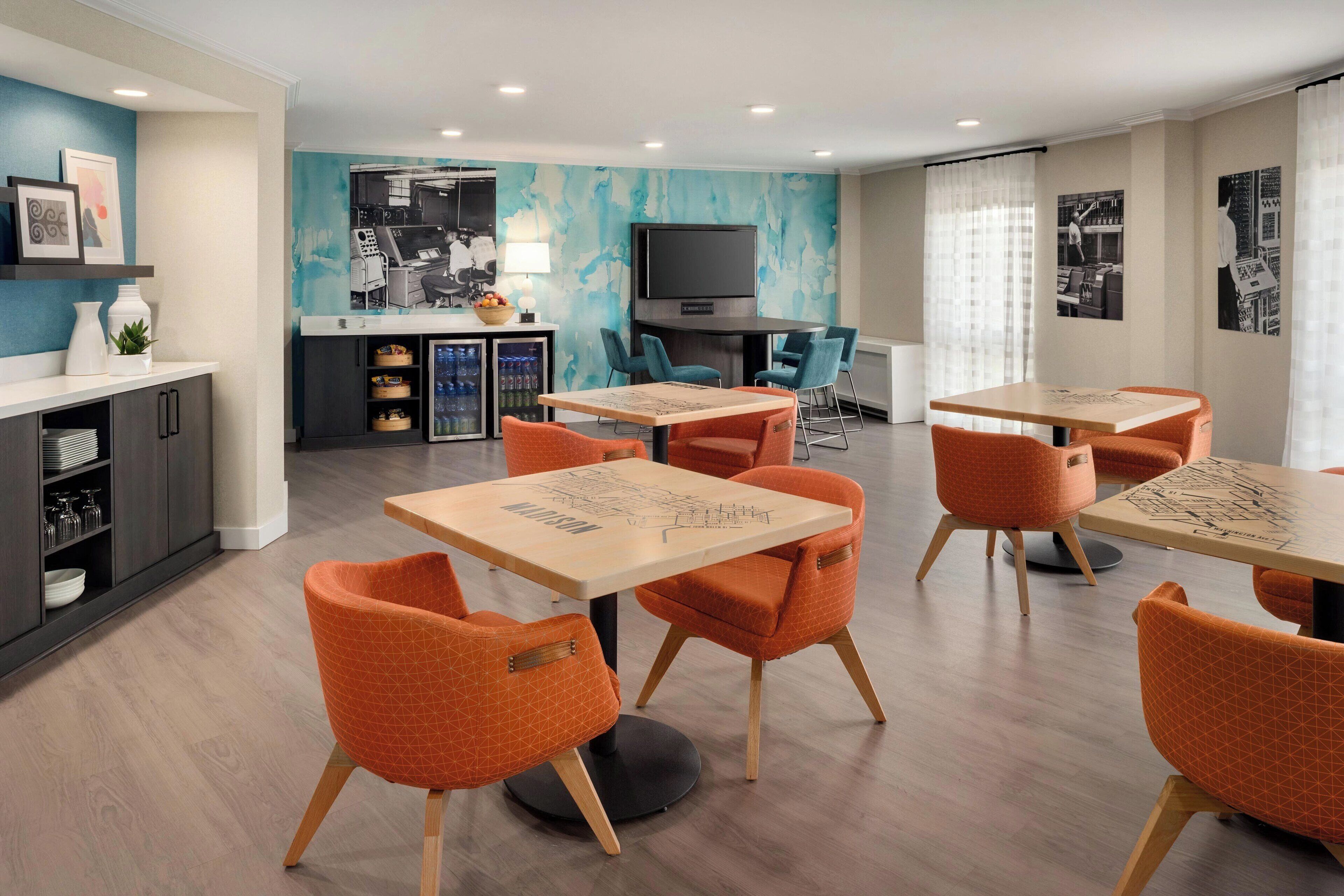 Un espace repas moderne avec des chaises orange et des tables en bois, un coin cuisine équipé d’un téléviseur, et un mur bleu orné d’œuvres d’art abstraites.