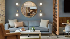 A modern living room with a grey sofa, a coffee table, and a round mirror. There are two wall-mounted light fixtures and a wooden ladder structure.