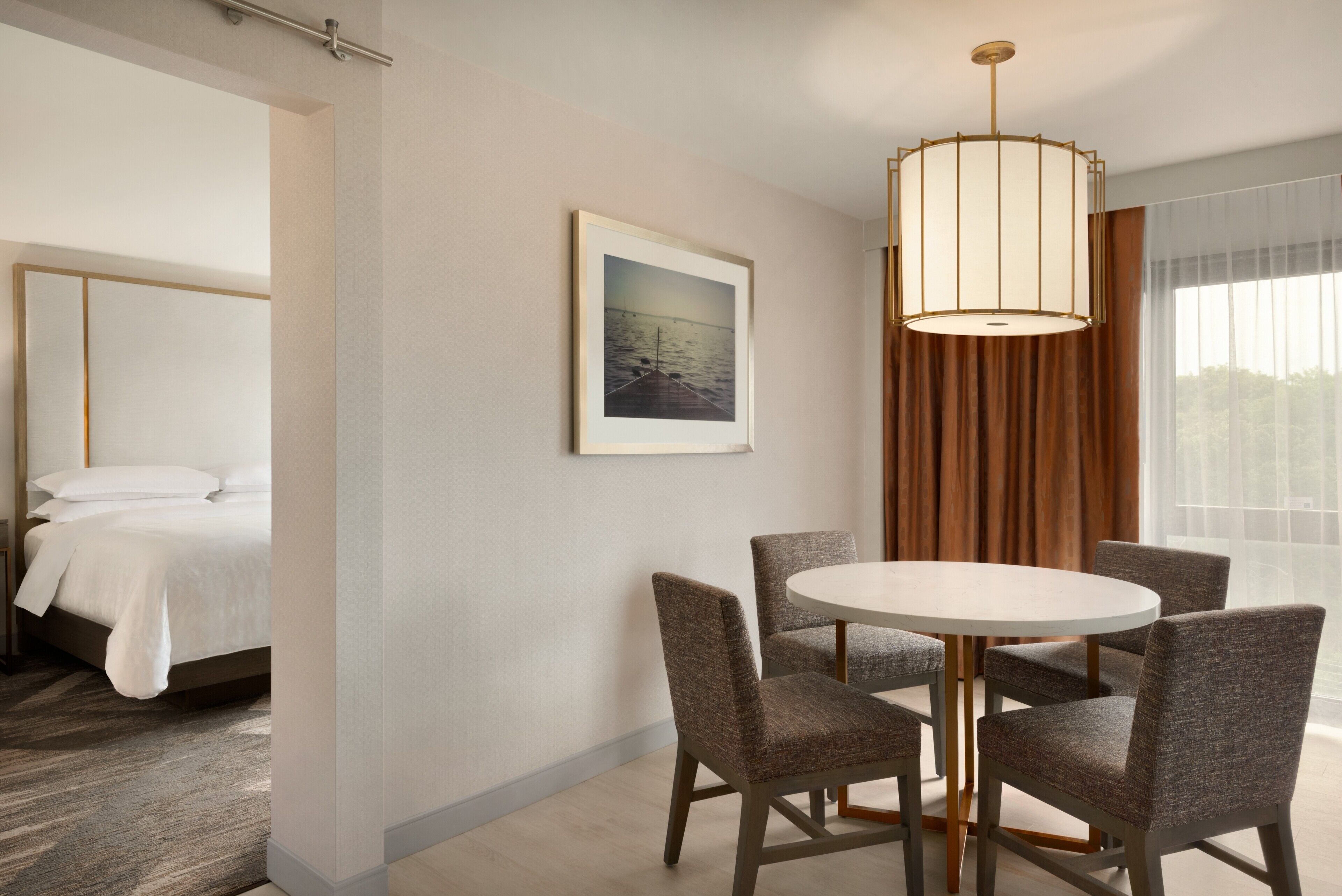 A hotel room with a bed, a dining table with chairs, a framed picture on the wall, and a large window with curtains.