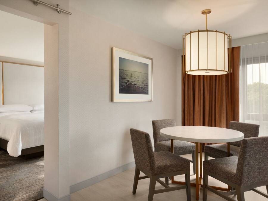 A hotel room with a bed, a dining table with chairs, a framed picture on the wall, and a large window with curtains.