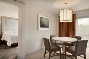 Habitación de hotel con cama, una mesa de comedor con sillas, un cuadro en la pared y un ventanal con cortinas.