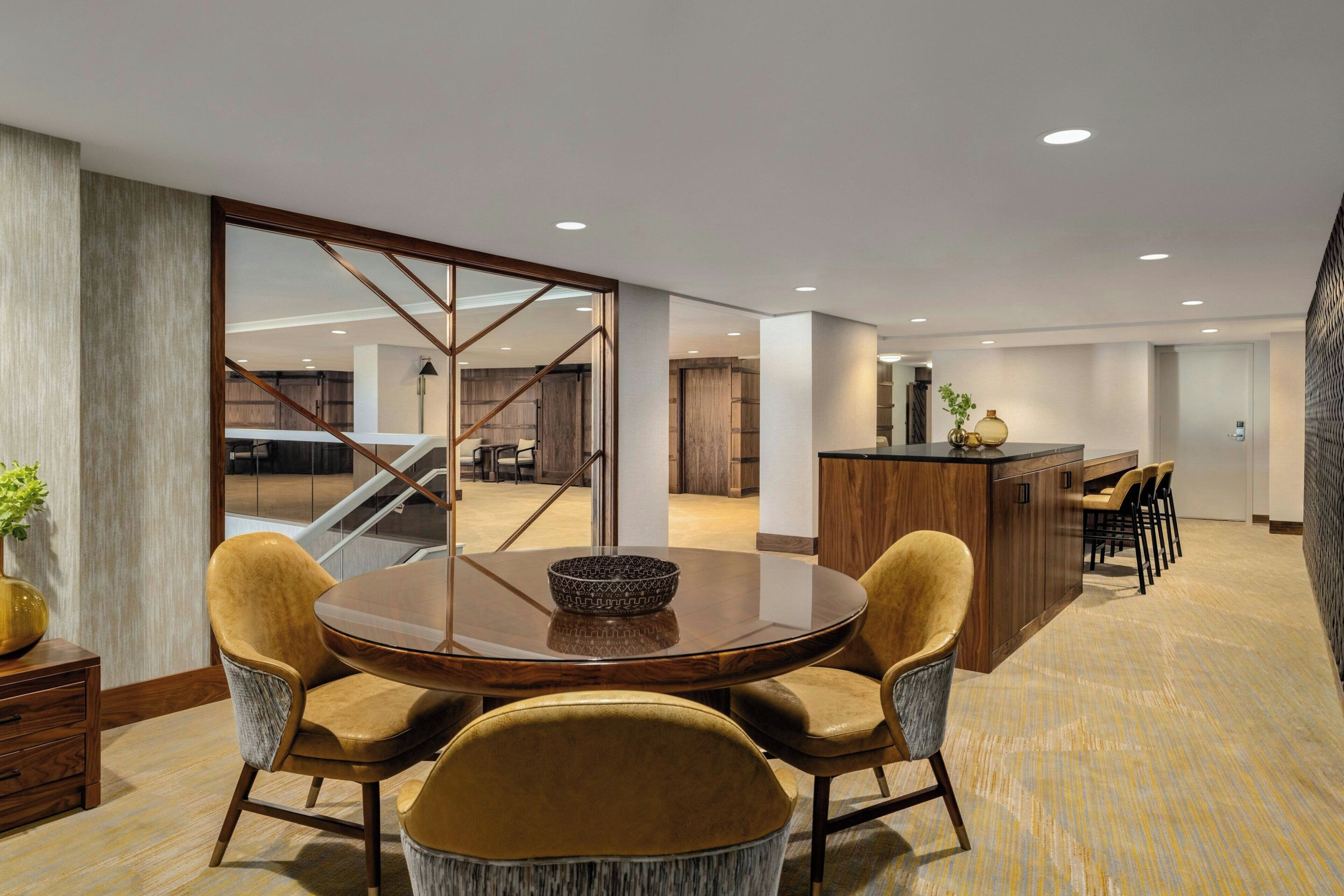 A modern hotel room with a dining area featuring a round table and four chairs, a kitchen area with a bar, and a view of the parking garage.