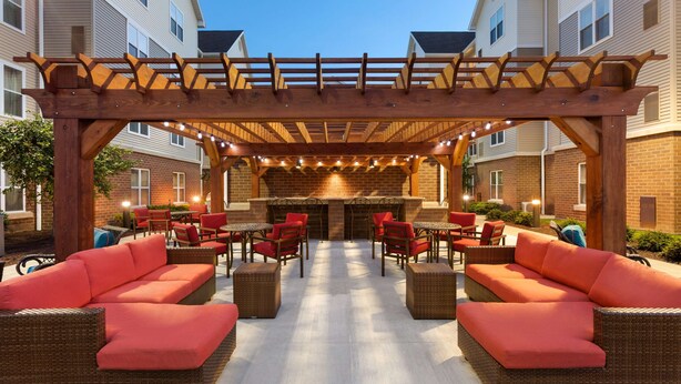 An outdoor patio area with wicker furniture and red cushions, a brick fireplace, and a pergola with string lights. - Homewood Suites by Hilton Harrisburg-West Hershey Area (Mechanicsburg)