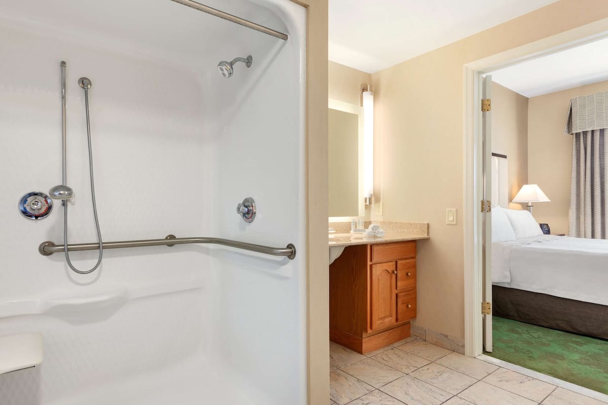 Une salle de bain d’hôtel avec une baignoire, une douche et des toilettes. On peut voir un meuble en bois et un lit à travers une porte ouverte.