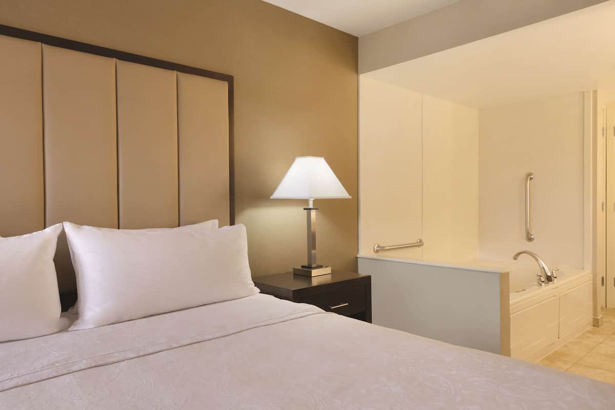 Une chambre d’hôtel avec un lit, une table de chevet avec une lampe, et une salle de bain avec un lavabo et une douche.